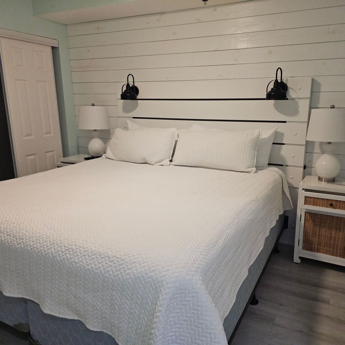 bedroom with white shiplap walls, wooden headboard on a king bed, nightstands and lamps on either side of the bed
