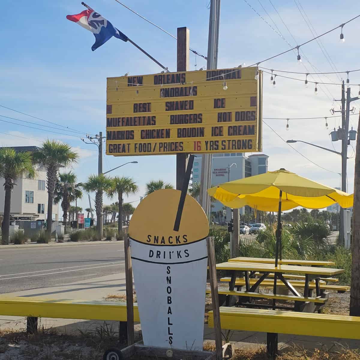 Yellow sign with New Orleans Original Snoballs, yellow and white snow cone sign, yellow picnic tables and umbrellas