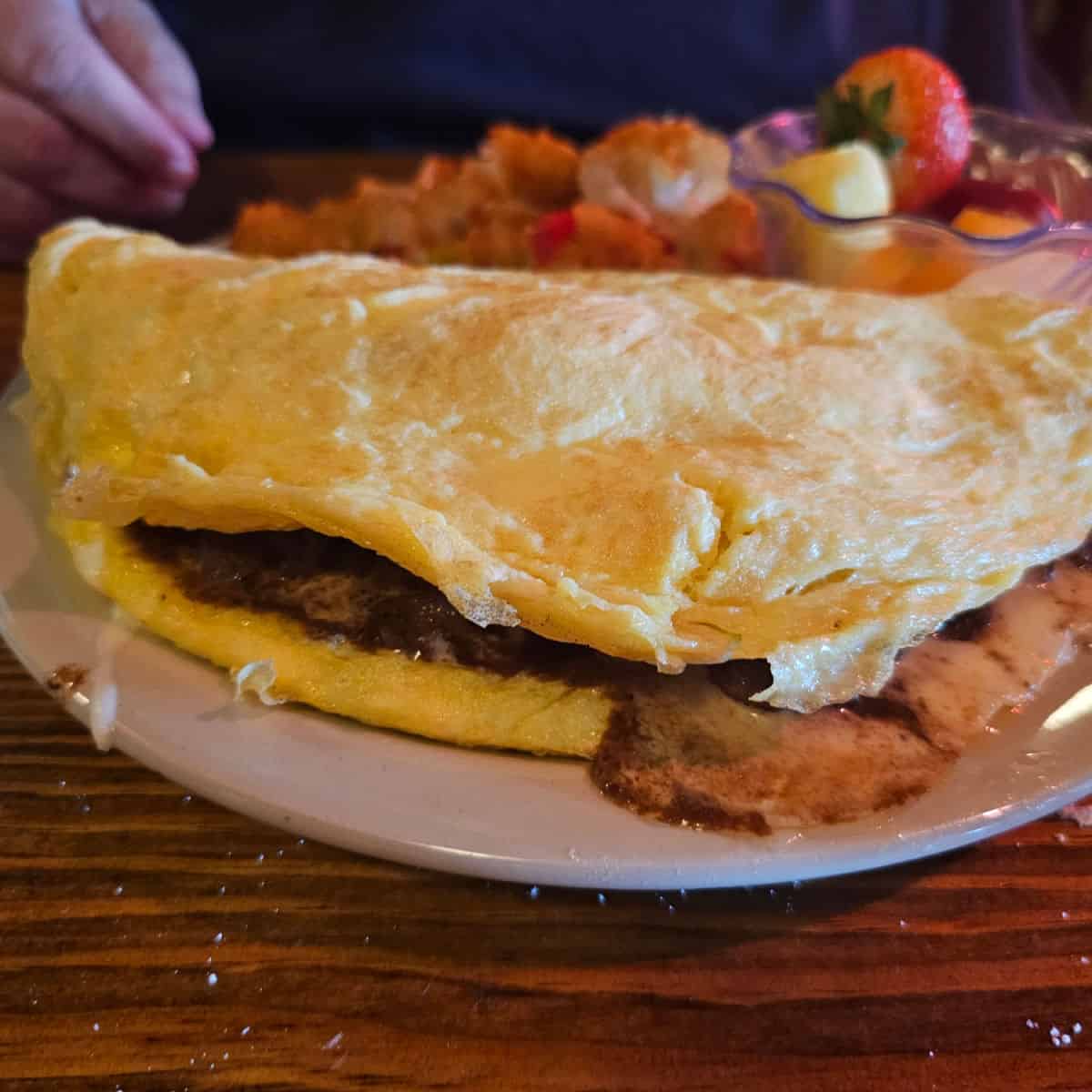 Steak omelet on a white plate with potatoes and a cup of fruit in the background