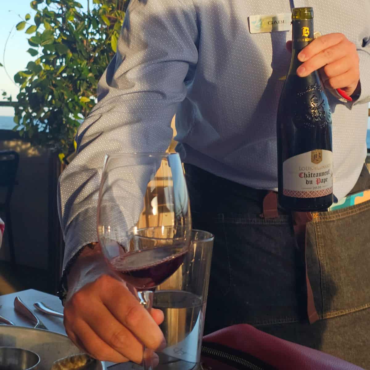 hand holding out a glass of wine while the other hand holds the bottle showing the label