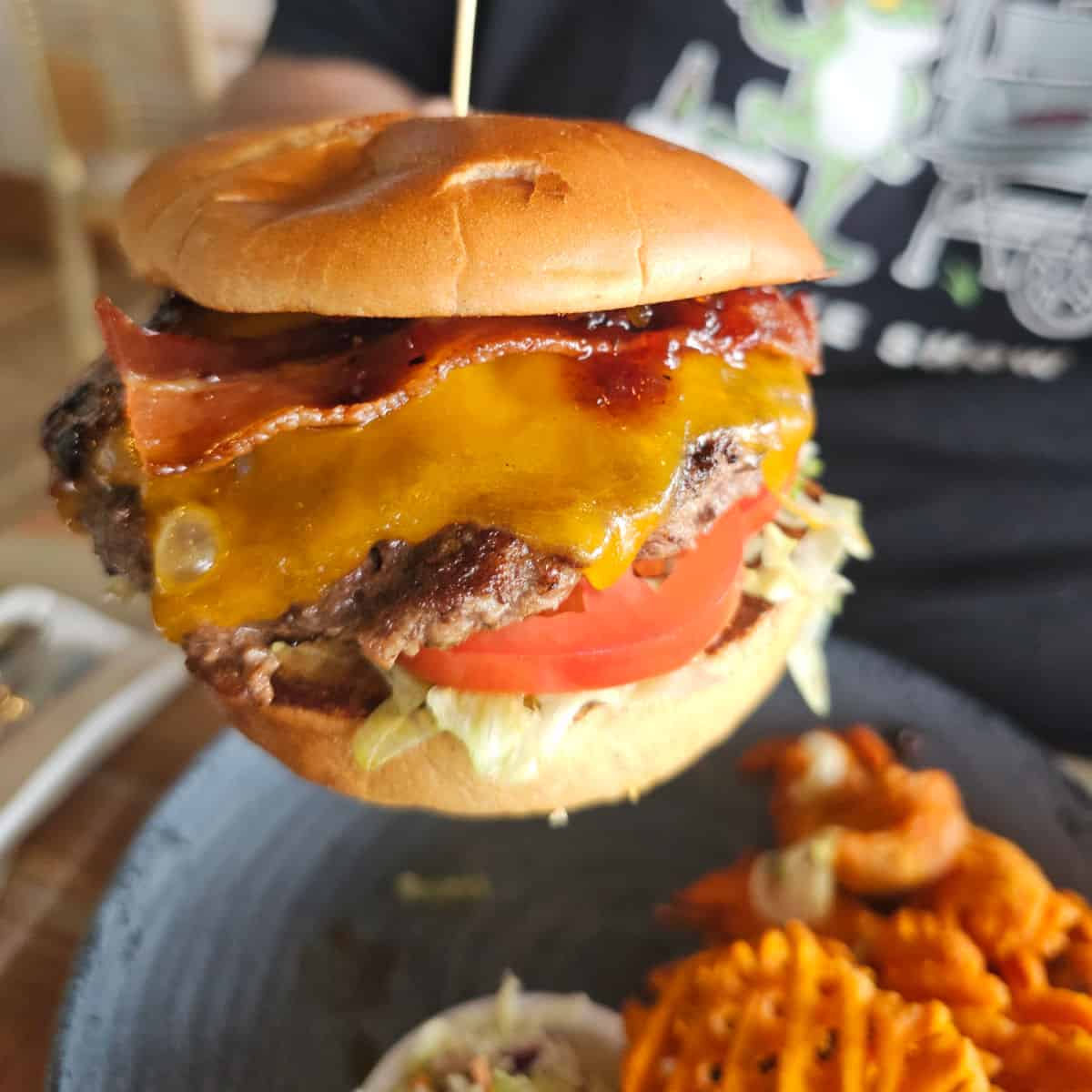 hand holding a hamburger with bacon and cheese over a plate with sweet potato fries