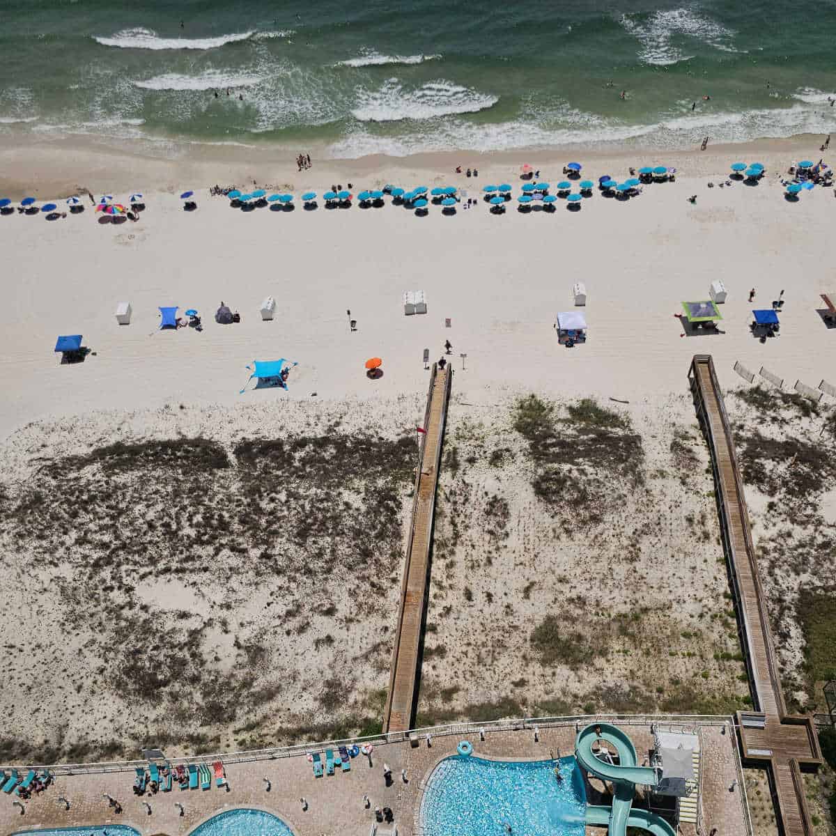 looking down for a high floor to a pool with waterslide, boardwalks leading out to the beach, colorful umbrellas on the beach and Gulf water