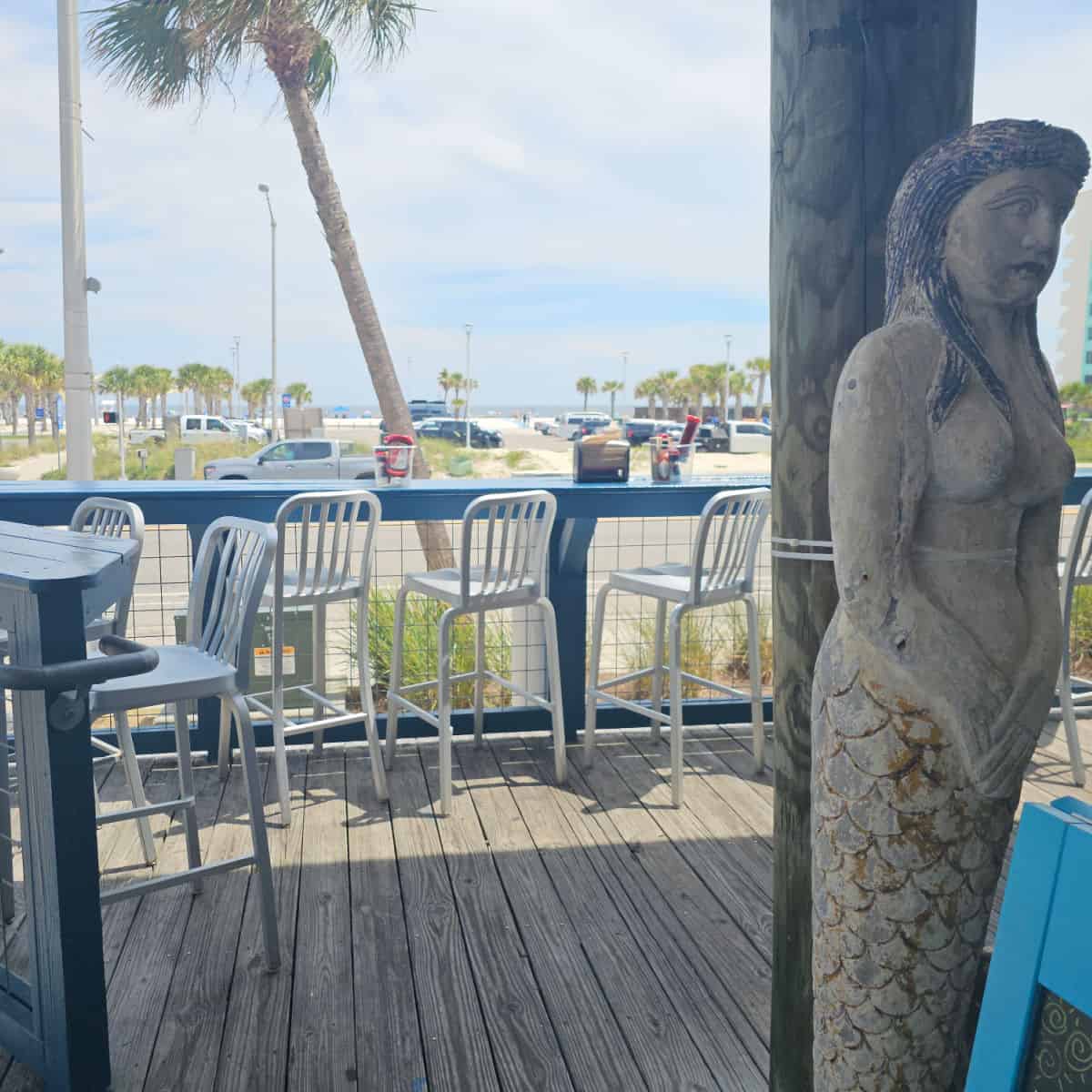 mermaid art against a pole looking out to chairs against a blue rail with palm trees and the public beach across the street