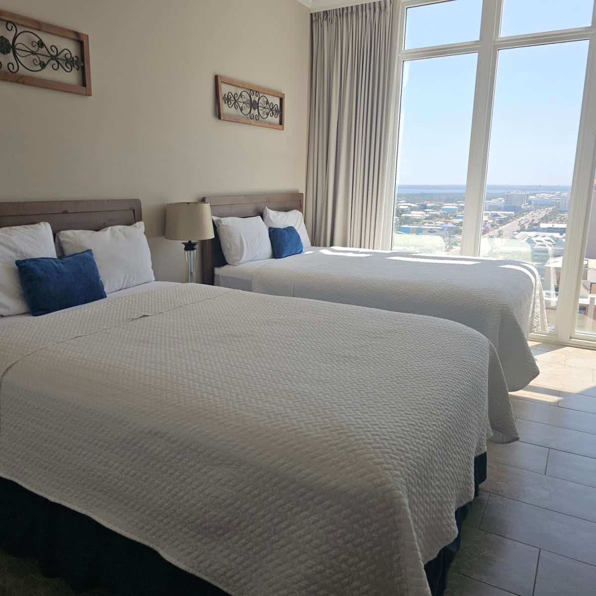 bedroom with two queen beds with white linens and a blue pillow next to a large window looking out over Gulf Shores