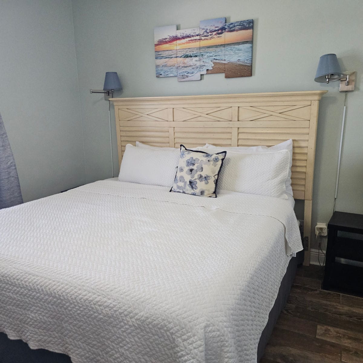 bedroom with wooden headboard behind the bed with white linens and a flower pillow, coastal beach art above the bed