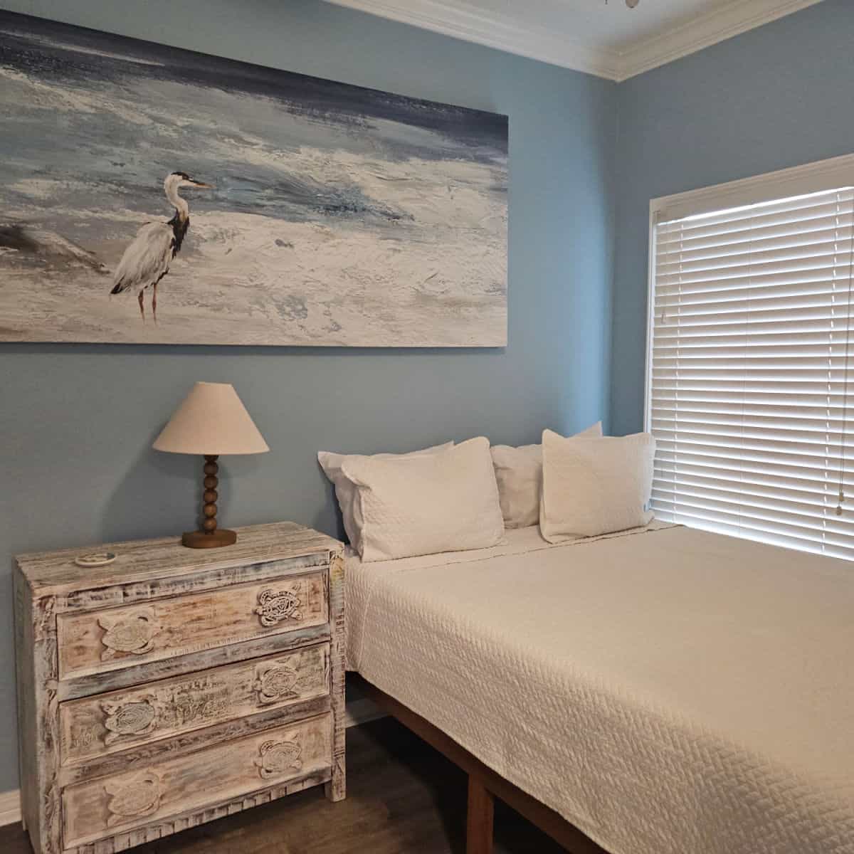 Bedroom with bed against the wall, blue heron art on the blue wall, cabinet with lamp and coaster
