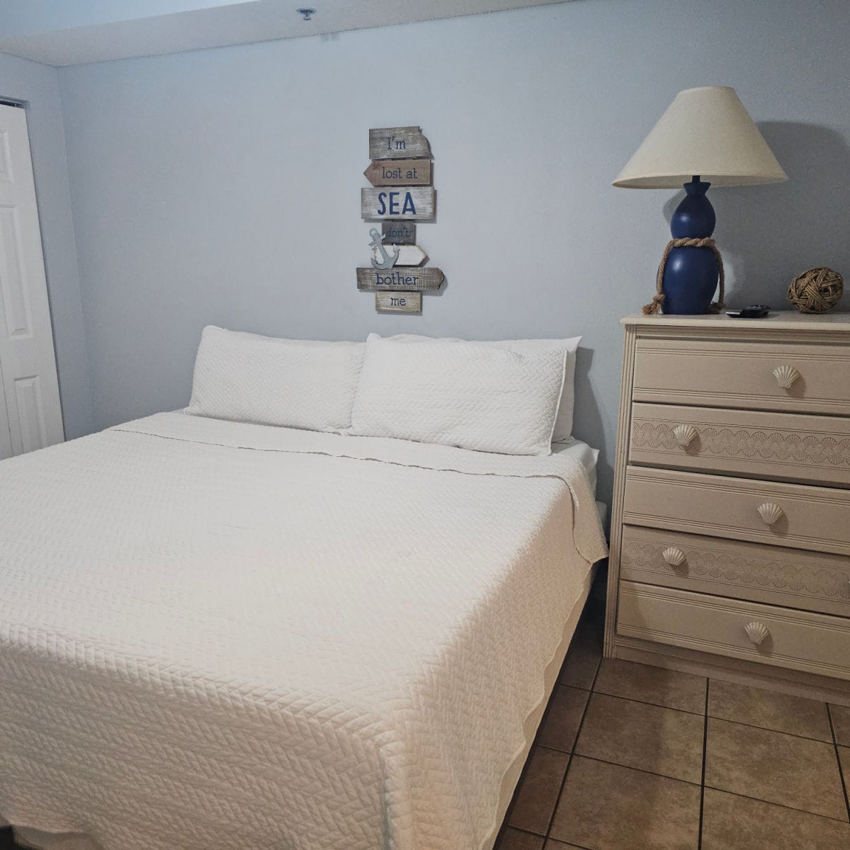 bedroom with blue walls, lost at sea art above a queen size bed with white linens, dresser with shell handles, blue lamp on the top of the dresser
