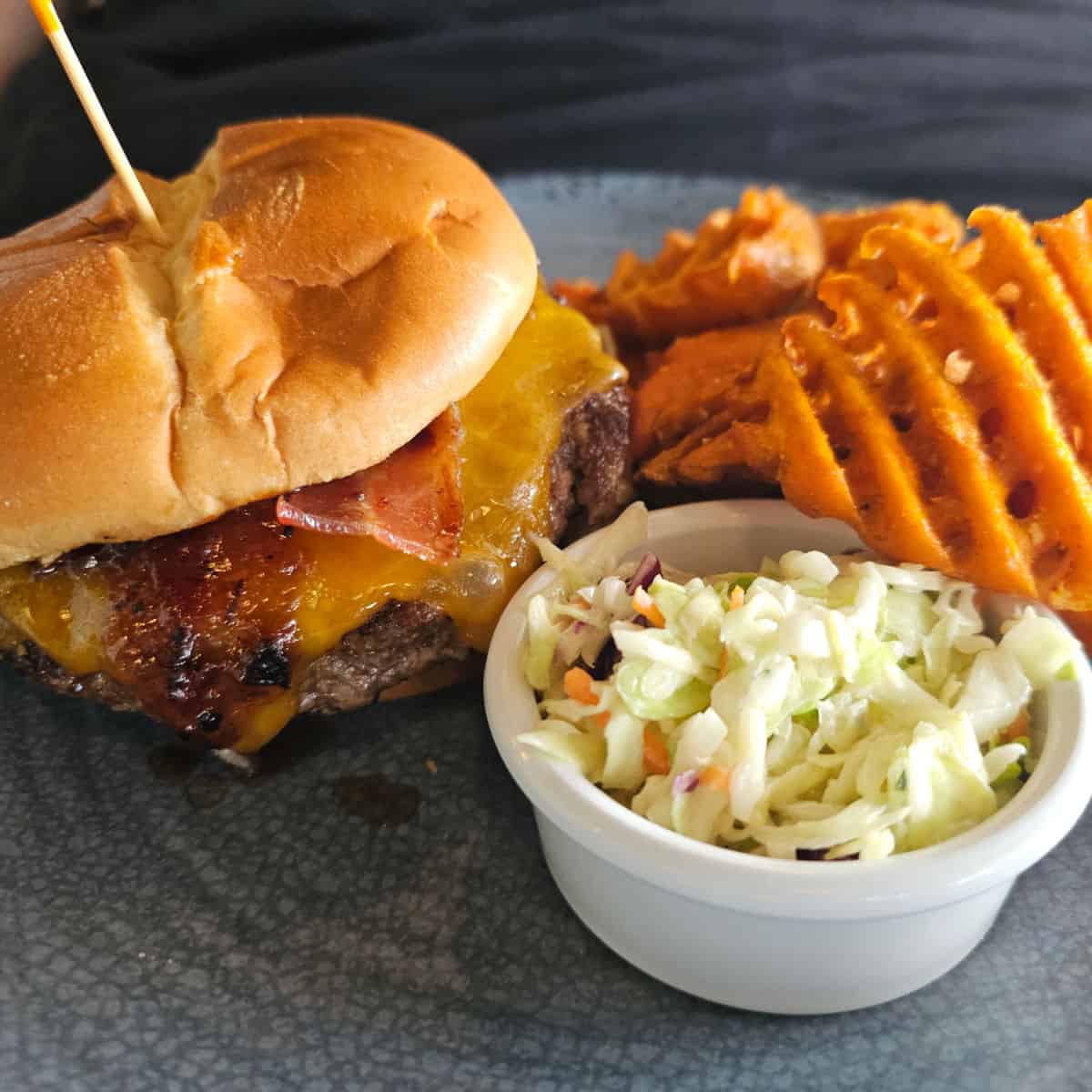 bacon cheeseburger and sweet potato fries with a container of coleslaw on a blue plate