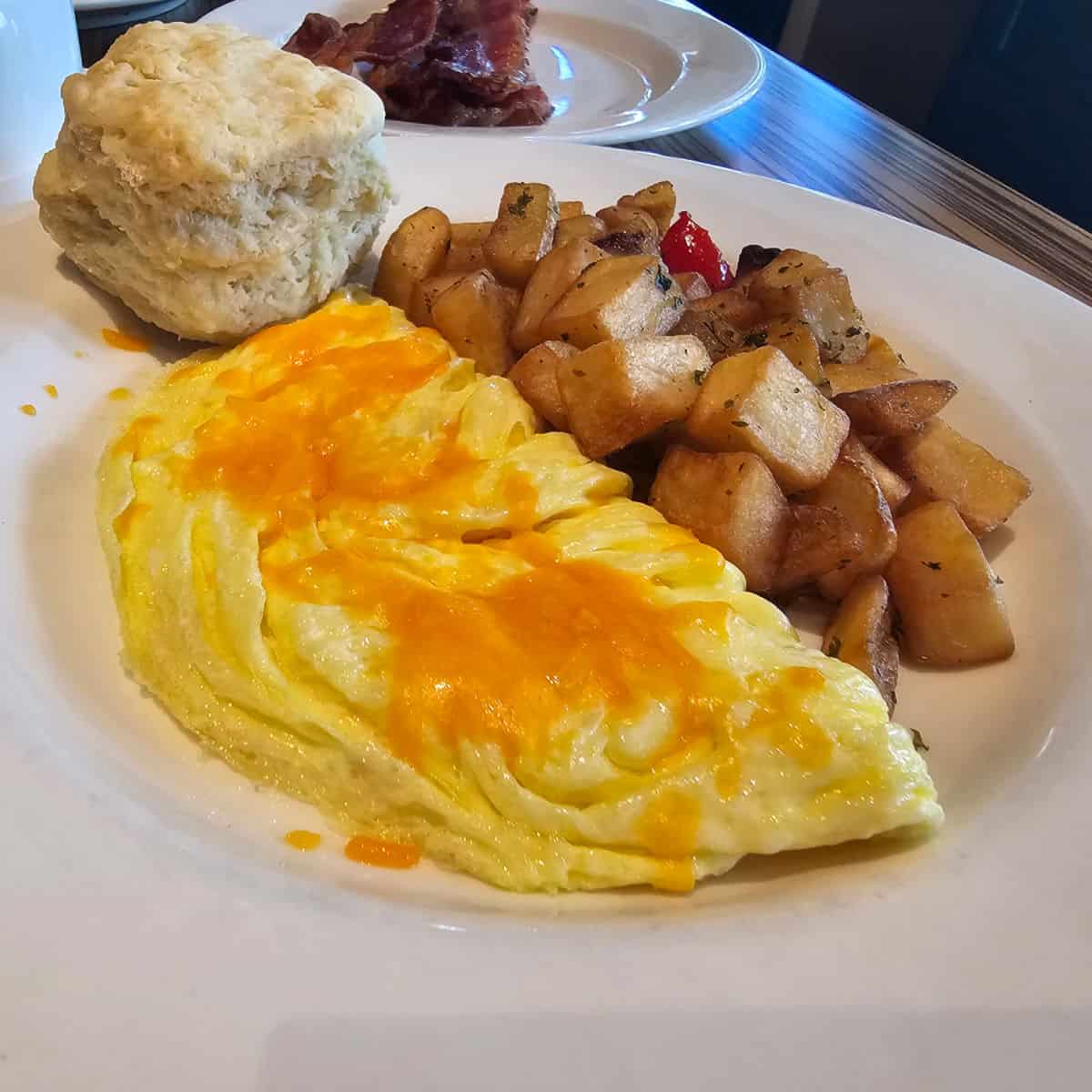 cheese omelet and home frie potatoes on a white plate with a biscuit, plate of bacon in the background
