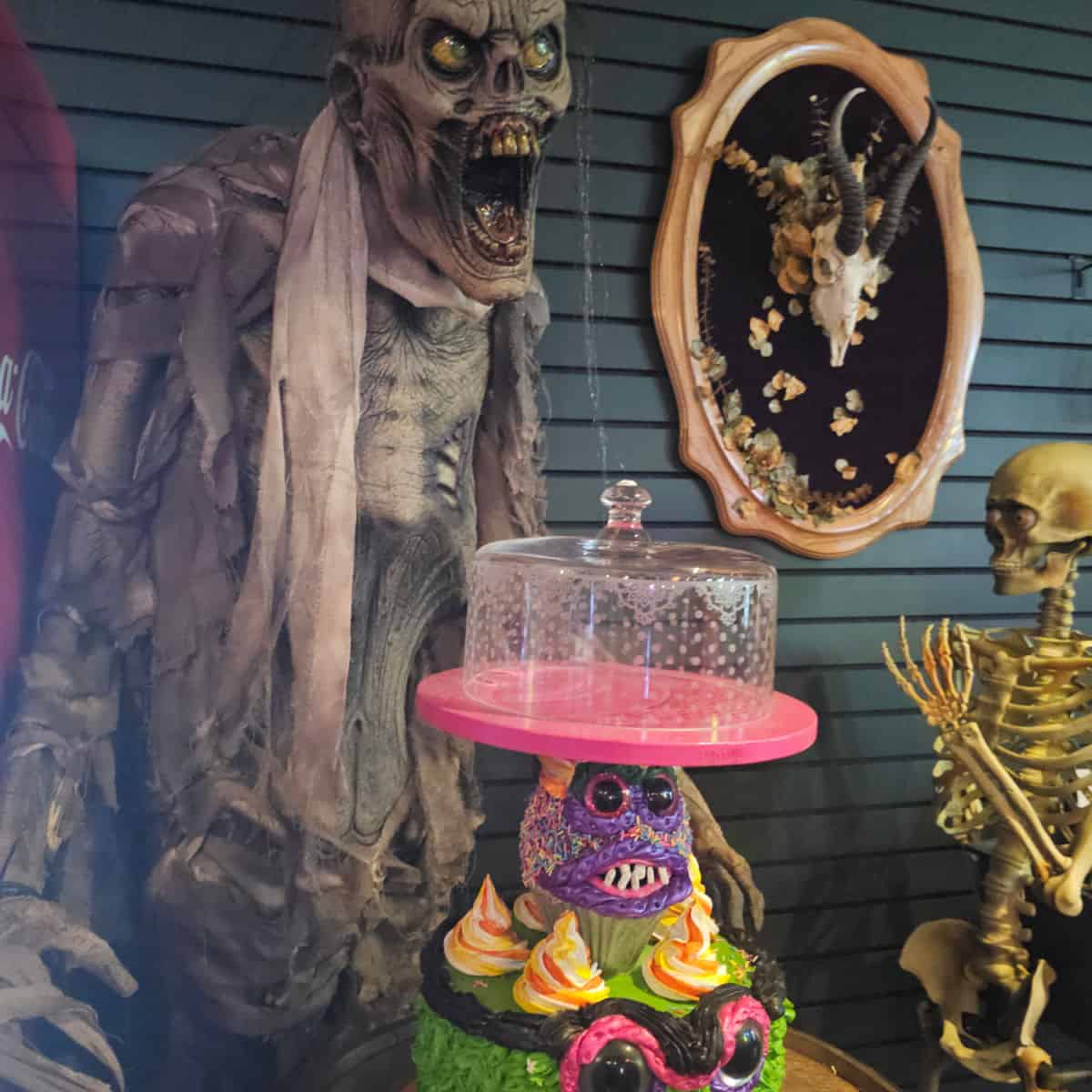 cake stand with a purple monster and cupcakes on it, with a large skull creature screaming behind it, gold skeleton to the side, horned skull on the wall