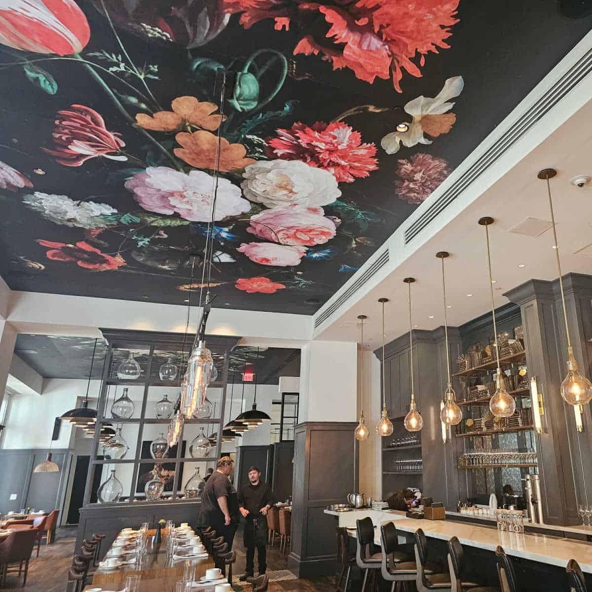 Ovide Restaurant interior with floral mural on the ceiling, pendant lamps over a long table, bar area to the right