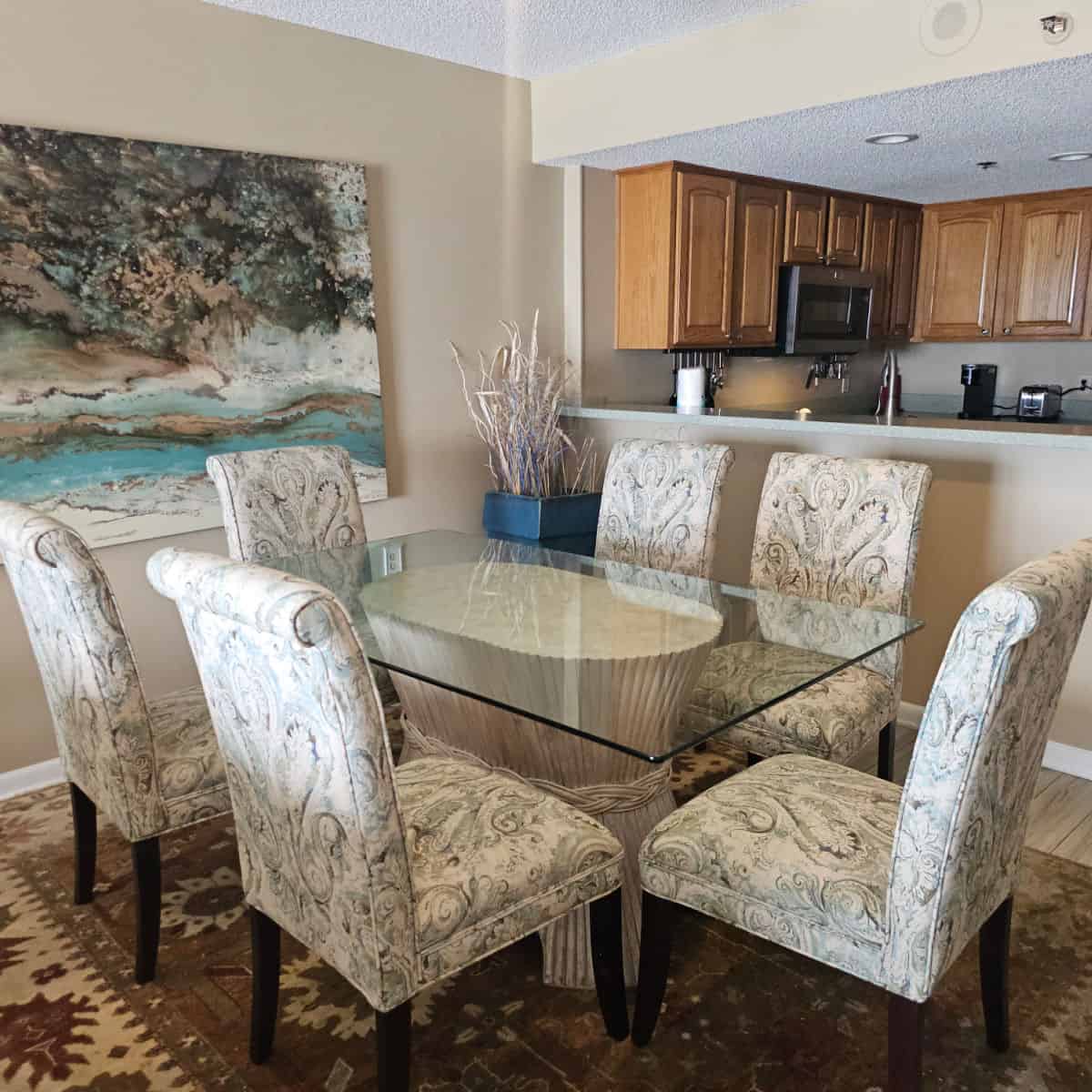 living room table with glass top, white paisley padded chairs around the table, looking towards the kitchen with wooden cabinets and stainless steel appliances