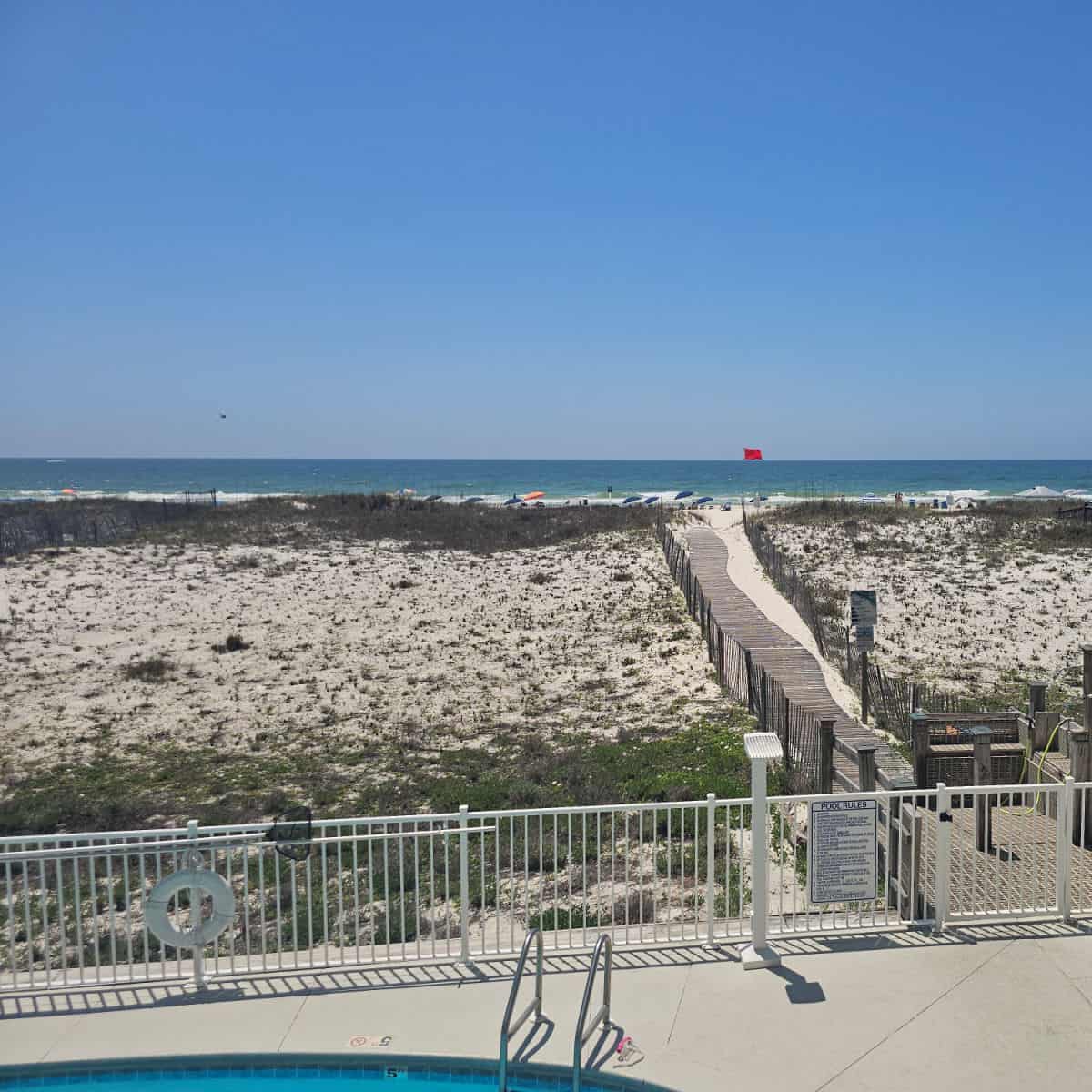 looking over a pool deck with a white fence leading to a walkway through the sand to the beach with a red flag, white sand and Gulf waters