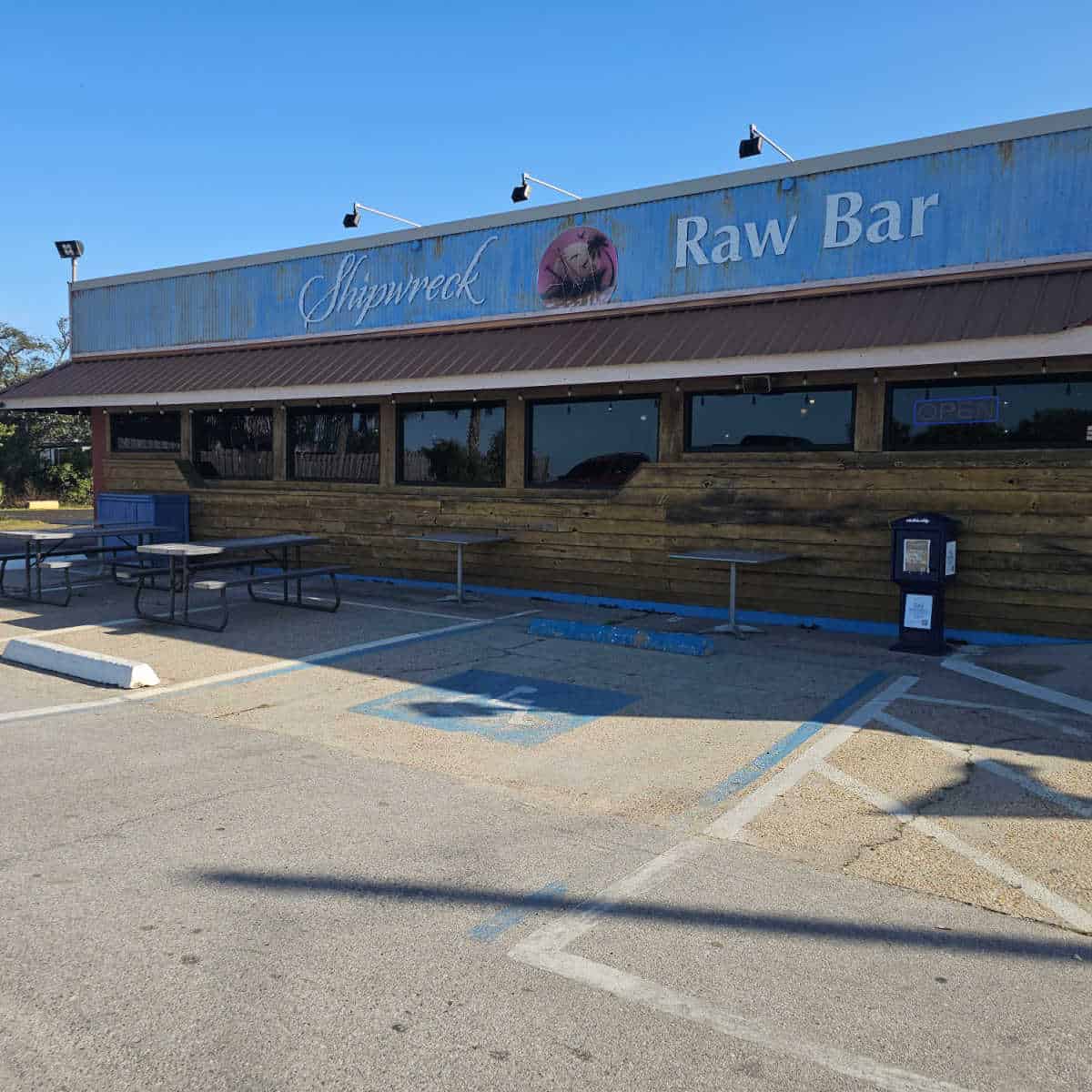 Shipwreck Raw Bar sign on a blue awning over the top of a wooden building, picnic tables in the parking area