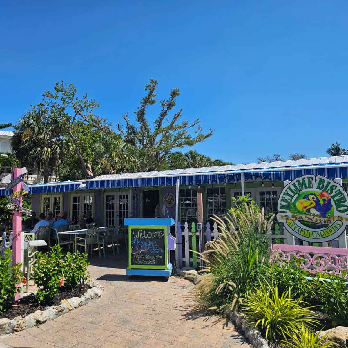 Key Lime Bistro sign with a Toucan on the right side, blue and white striped awning on the restaurant with a welcome sign at the hostess desk, tables and chairs in the background