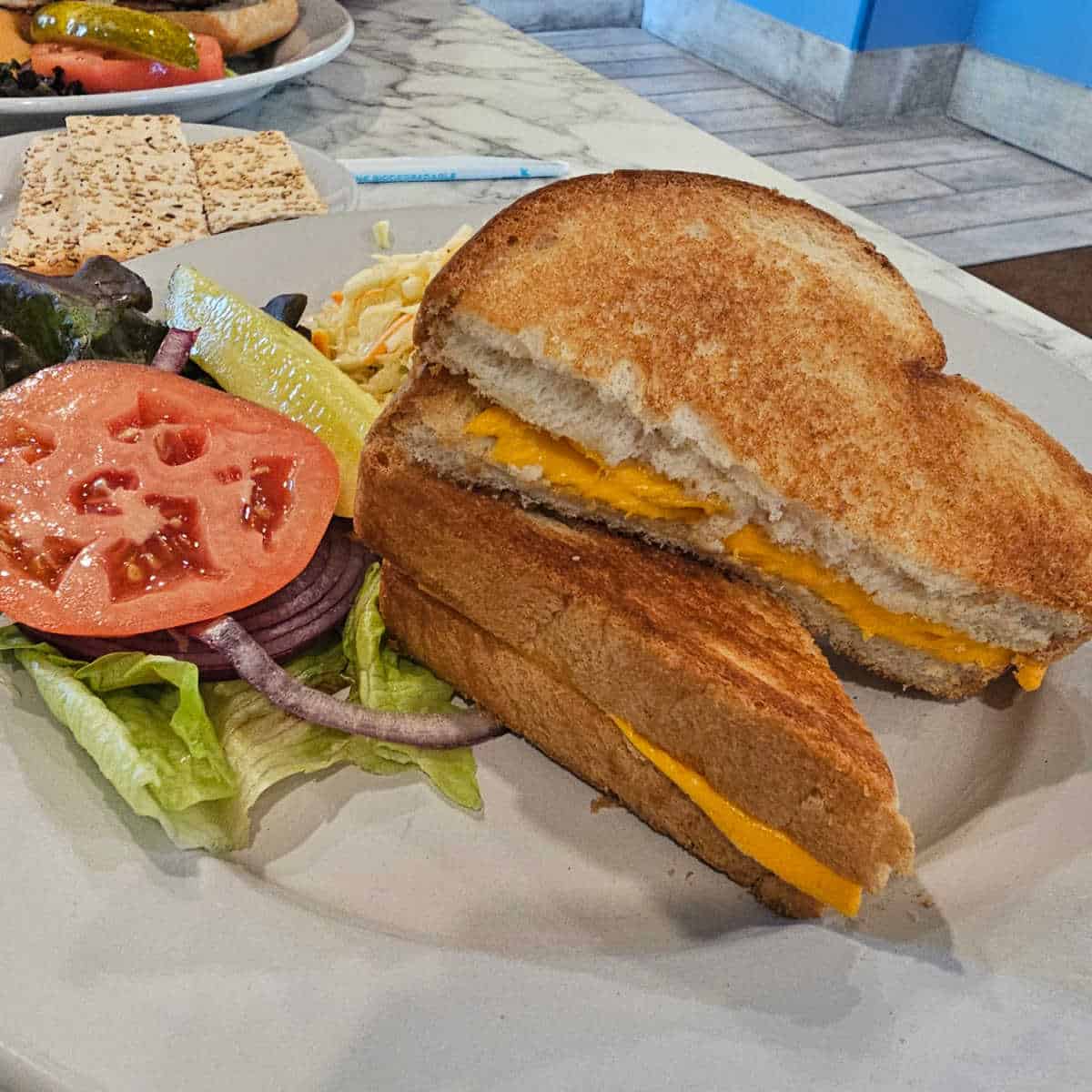 grilled cheese sandwich on a white plate with crackers near it and tomato and lettuce