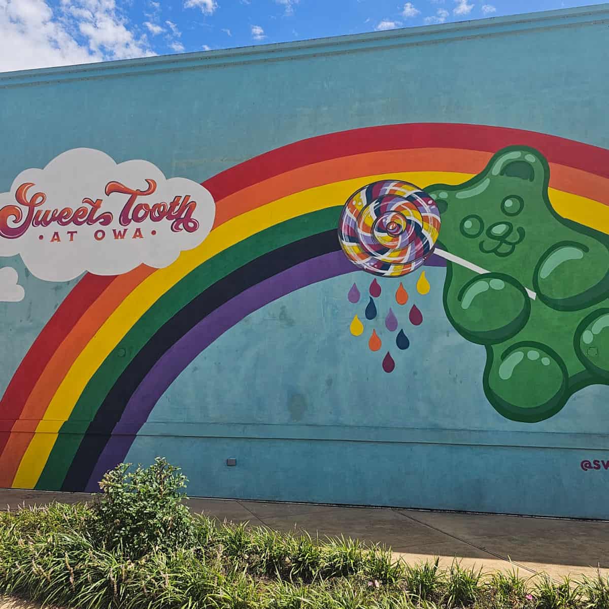 Sweet tooth at OWA printed in a white cloud on a rainbow mural with a green gummy bear holding a lollipop