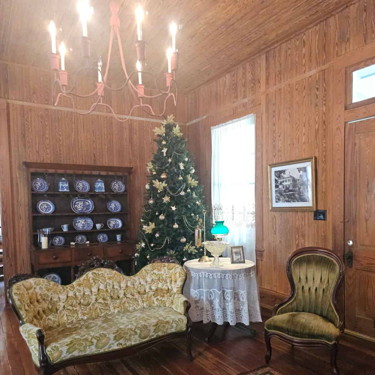 wood paneled room with a Christmas tree decorated in the corner, next to a hutch with blue and white plates, couch next to a white lace covered table with a lamp and glass bowl on a pedestal, brown and green chair by a door