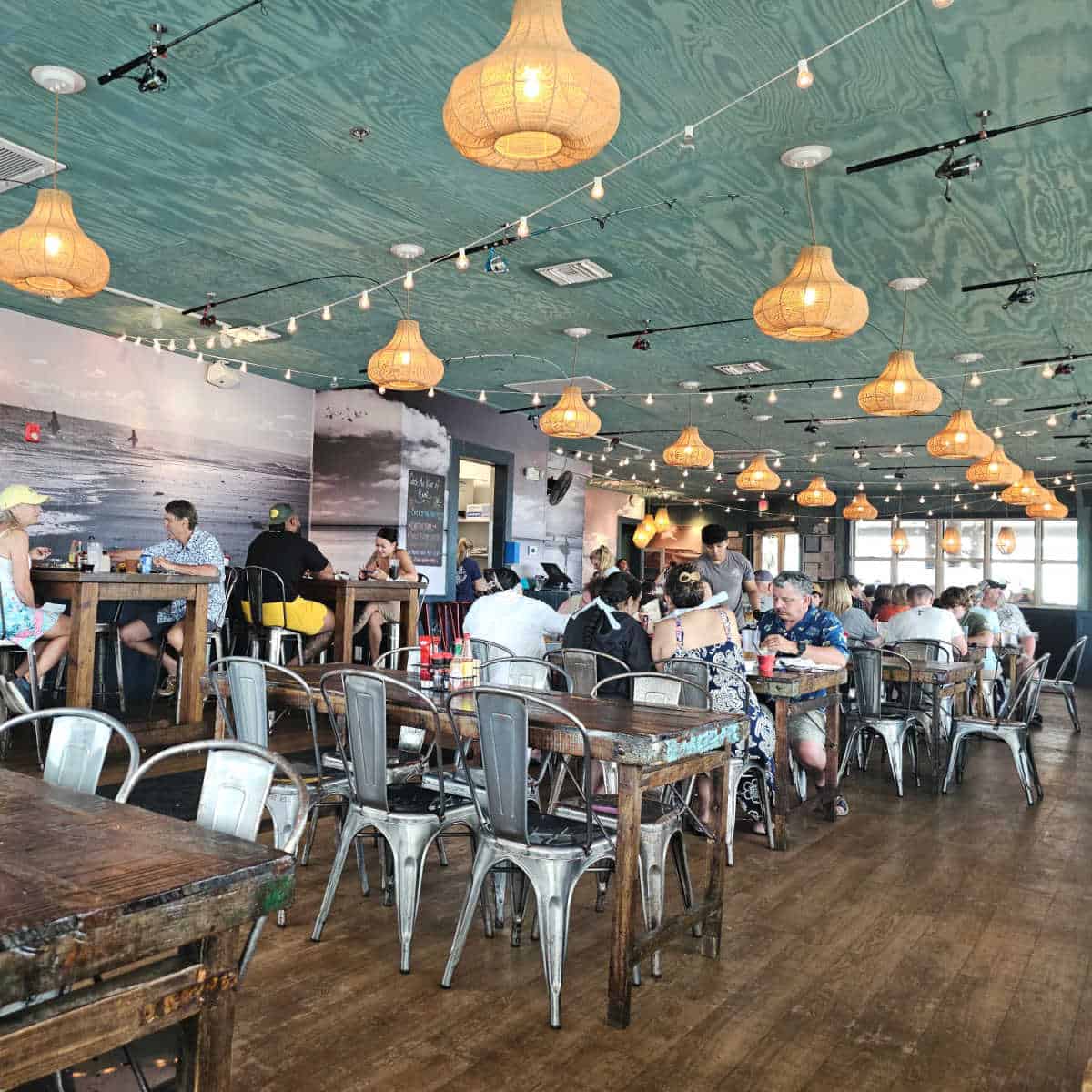 indoor restaurant seating with fishing poles holding the light shades, tables and metals chairs with diners spread throughout the restaurant, surf images on the wall