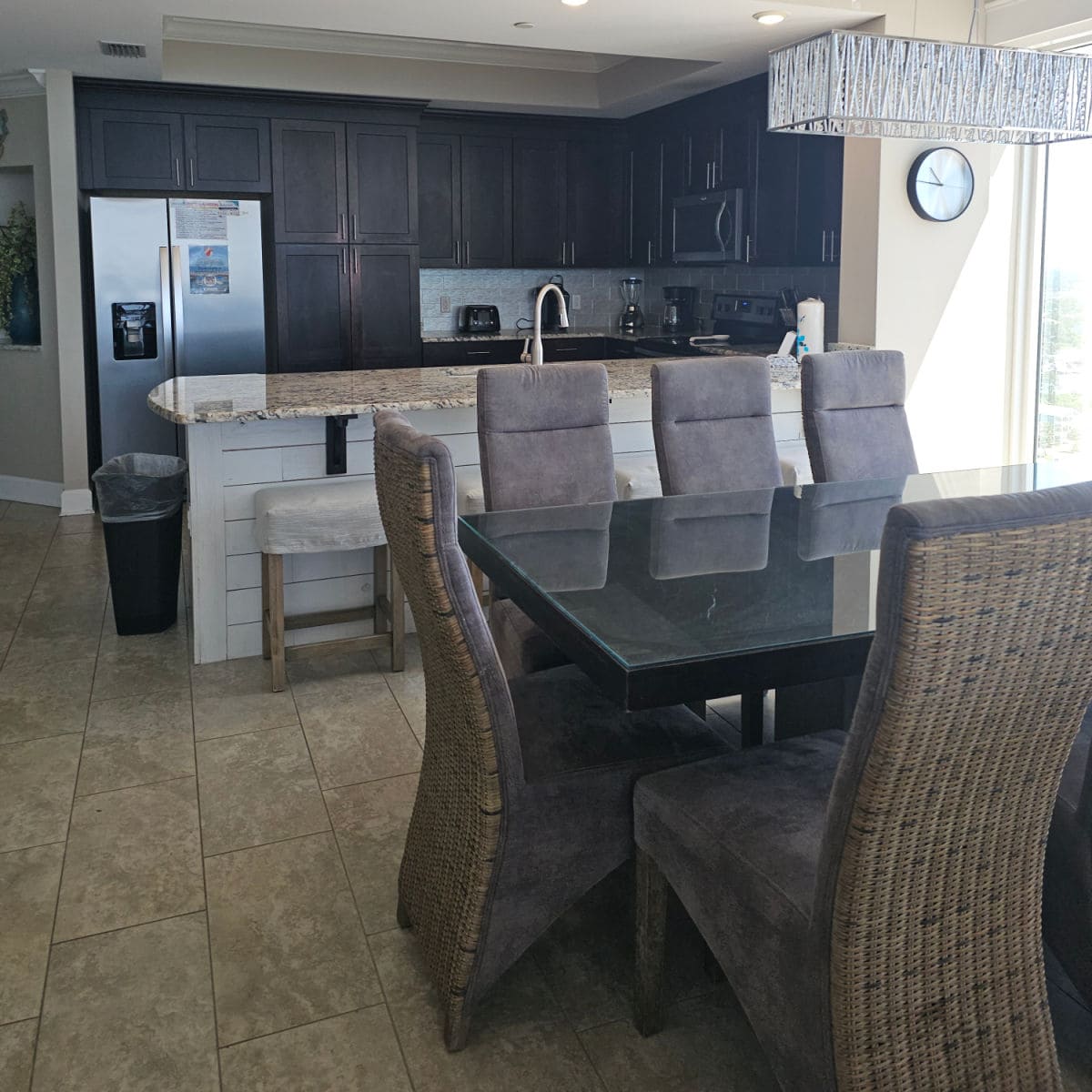 Kitchen and dining room area, table with padded grey chairs around the square table, kitchen with dark brown cabinets and stainless steel appliances