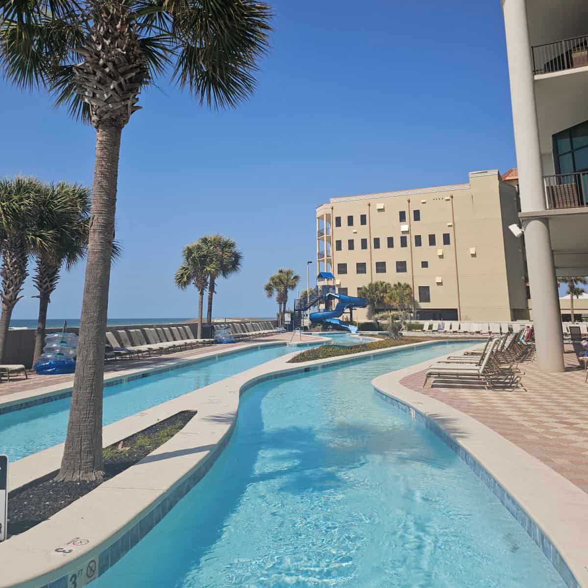 looking down a lazy river towards a waterslide, palm trees around the lazy river, lounge chairs on the pool deck