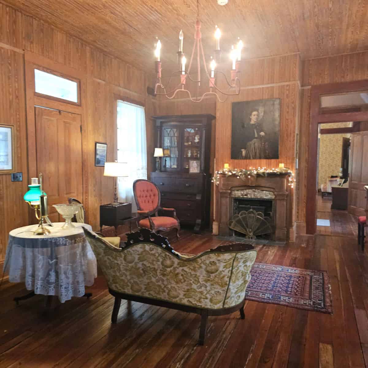 historic room with wood walls, antique cabinet on one side of a fireplace with a historic picture of a woman above it, small couch with green leaves on it next to a lace covered table with a lamp and glass bowl on a pedestal