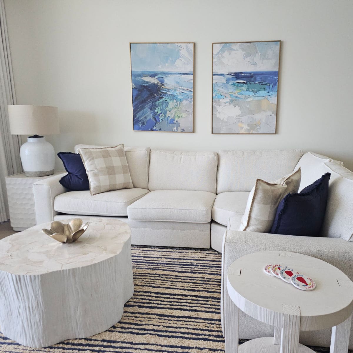 living room with white wrap around couch with blue and ginham pillows, blue and white art on the wall, white tables with beachball properties coasters