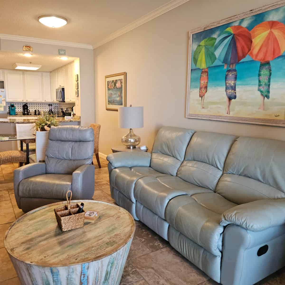 living room with a leather couch and recliner chair, circle table in the middle, wall art with three women holding bright umbrellas. Looking back towards the kitchen with white cabinets and stainless steel appliances