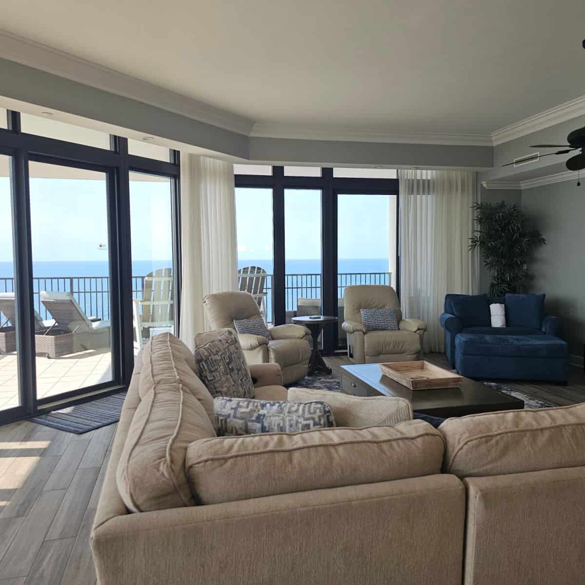 Living room with large wrap around couch, comfy blue chair, and two recliners. large windows looking out to a balcony with lounge chairs and Adirondack Chairs, gulf waters in the distance