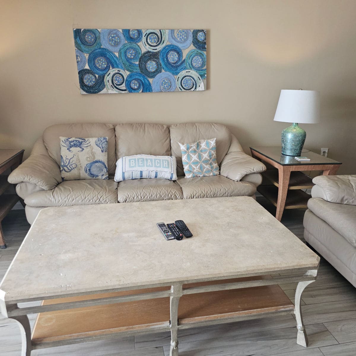 living room with leather couches, large table with the remotes on top of it, corner table with lamp on it, blue circular swirl art piece on the tan walls