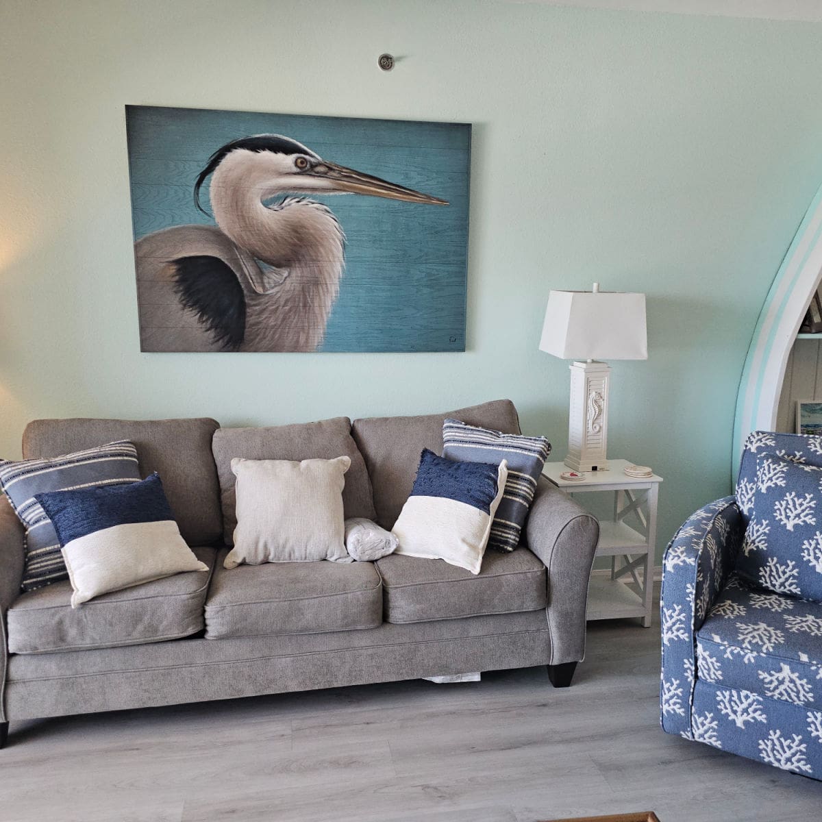 living room with an amazing Great Blue Heron art piece on the wall above a grey couch with pillows, blue chair with coral print