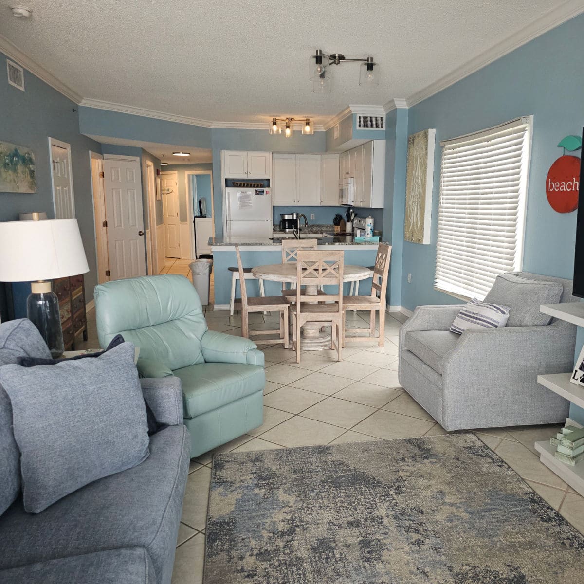 Living room with blue couch, grey and green recliners, looking towards the kitchen with 4 chairs around a table, white cabinet and appliances in the kitchen,