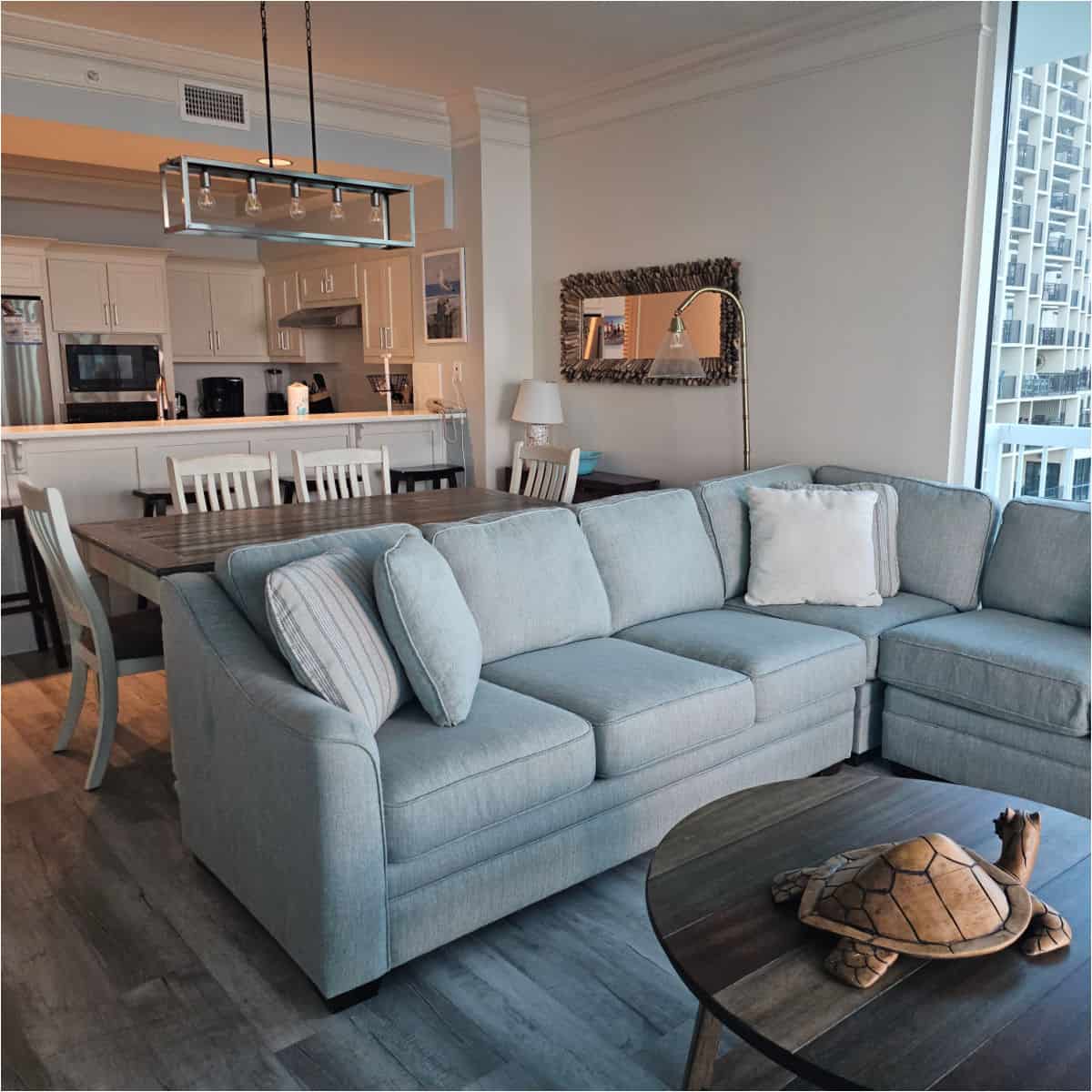 grey wrap around couch with pillows with a wooden table with wood turtle sculpture, table and cahirs with a light over it, looking towards the kitchen with white cabinets
