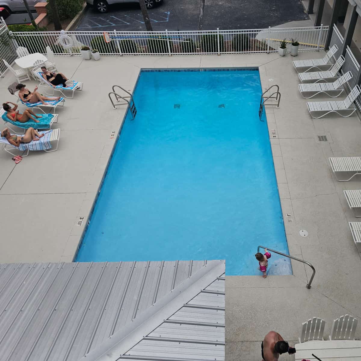 looking down over a rectangular pool with a kid in the water, lounge chairs around the pool deck, white fence around the pool