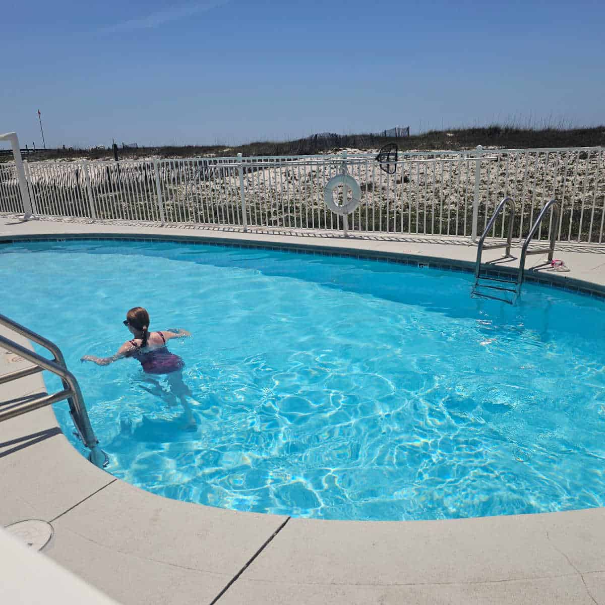 outdoor pool with a female swimming in the water, white fence with sand dunes behind it