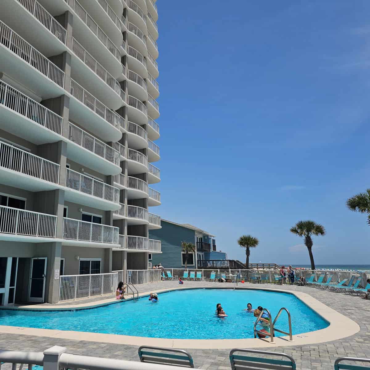 outdoor pool with people in the water, lounge chairs around the pool deck, tradewinds building next to the pool
