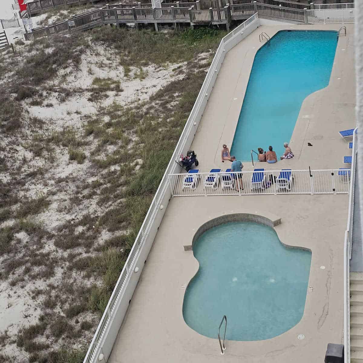 looking down over a pool with people on the edge of the pool, blue lounge chairs next to a fence, smaller pool with a ledge, all surrounded by a white fence. Sand dunes and walk way to the beach on the left side