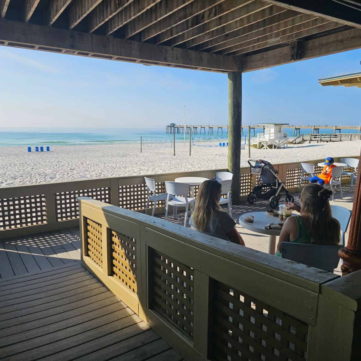 covered outdoor seating with white tables and chairs, a couple of people sitting in the corner looking out towards the beach and Gulf waters with a pier to the right side