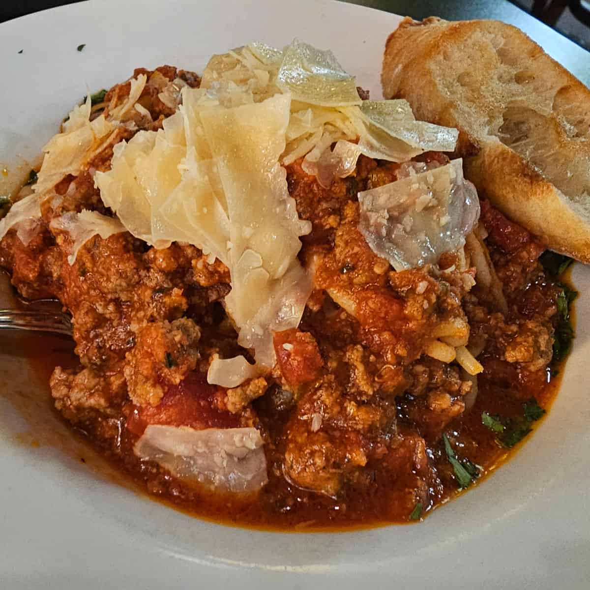 pasta with meat sauce and parmesan cheese on top next to a slice of bread in a white bowl
