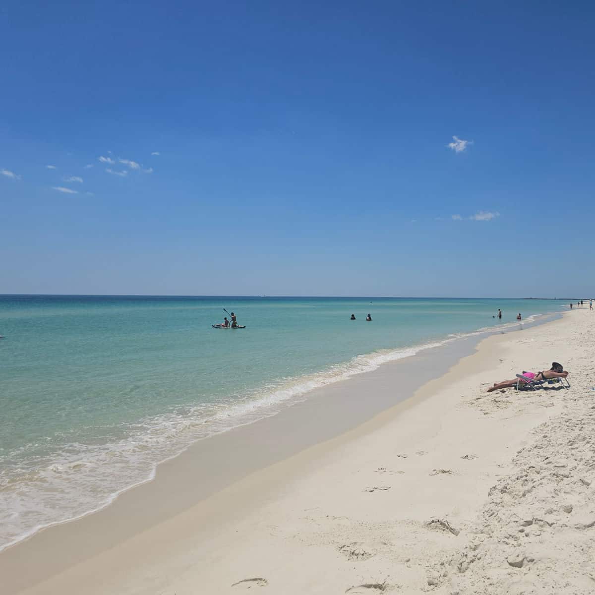 blue sky day with gorgeous turqouise Gulf water, people on a paddleboard and in the water, person lying on the white sandy beach