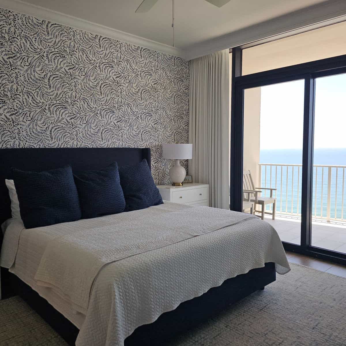Gulf facing bedroom with a large bed with white linens and large blue pillows, white nightstand with lamp, blue and white wall paper on the wall