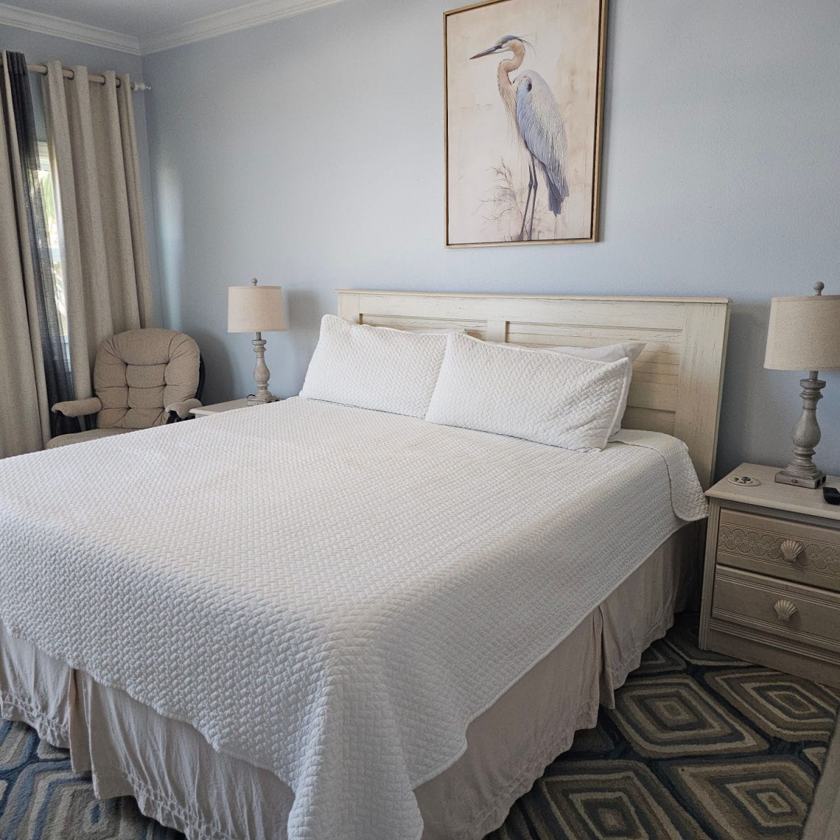 Bedroom with large bed with wooden bed frame, great blue heron art above the bed, chair in the corner, nightstand with a lamp next to the bed
