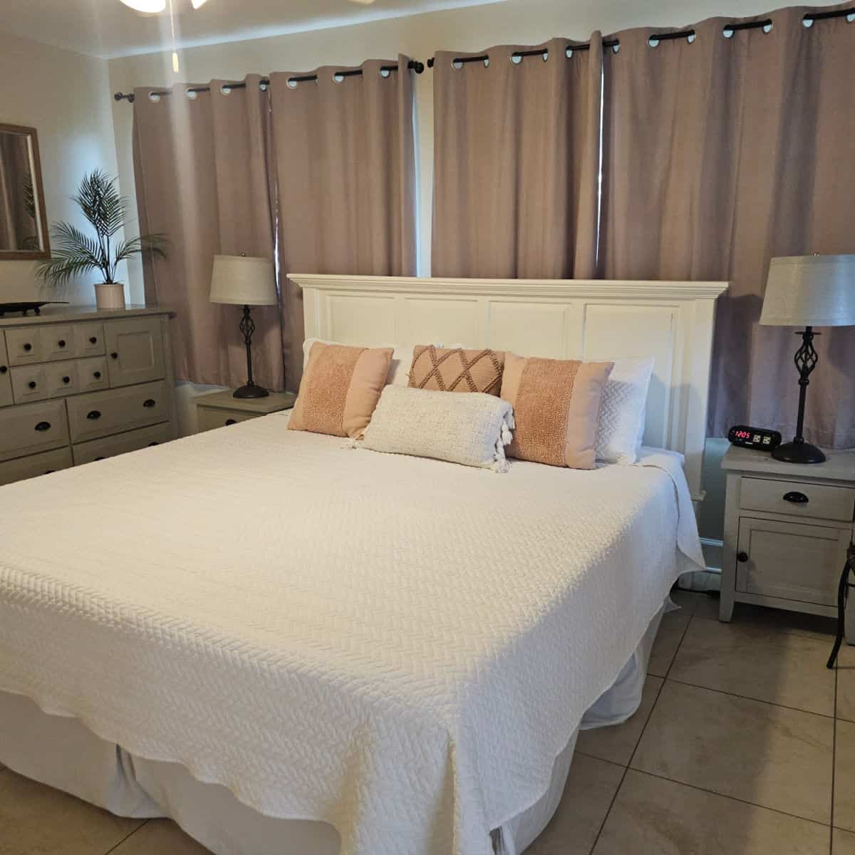 Large bedroom with king-size bed, tan and white pillows, two nightstands with lamps, a dresser along the wall with a fake plant on it. Tall curtains hanging behind the bed