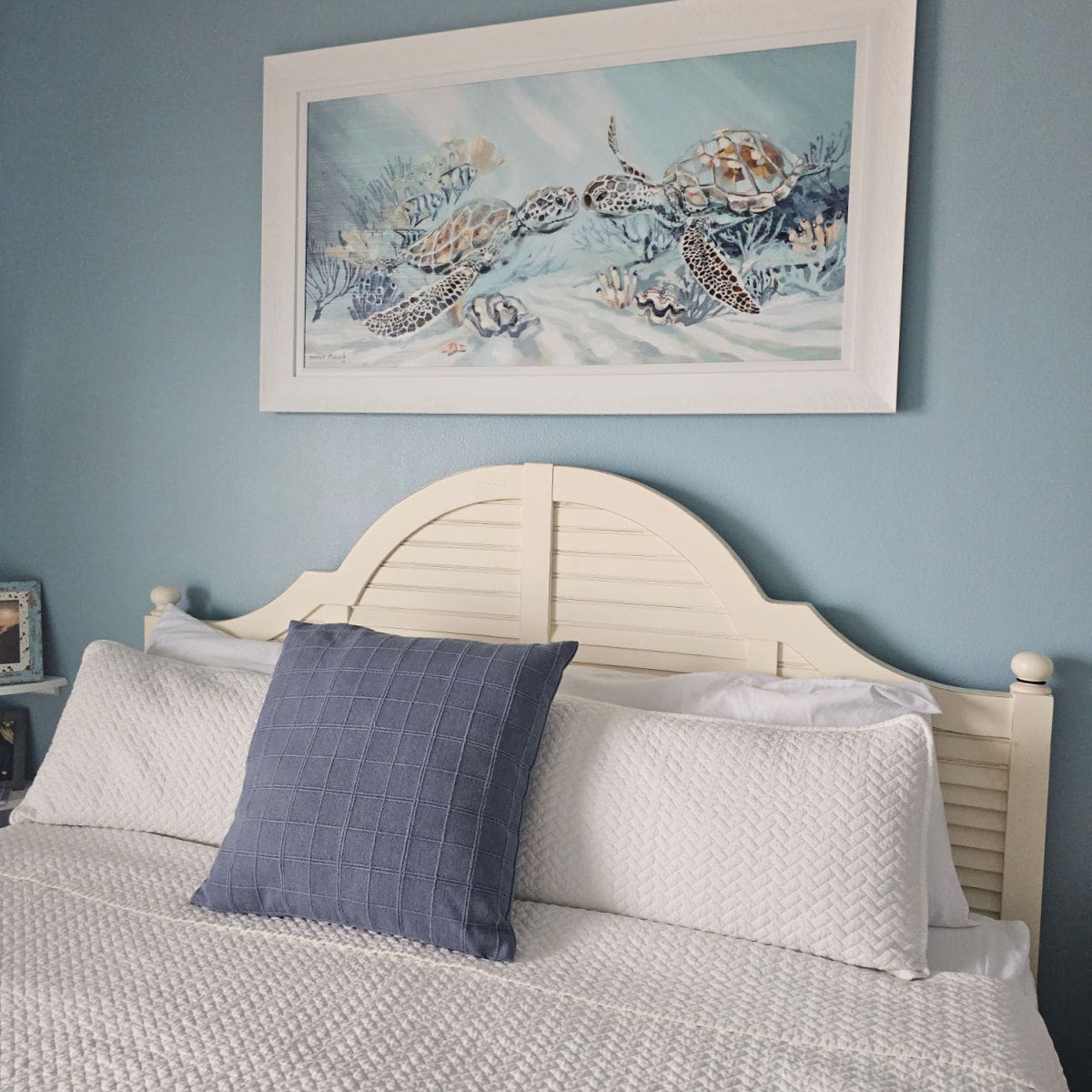 bedroom with blue walls, turtle print on the wall in a white frame, white wooden headboard with white and blue pillows, white linens