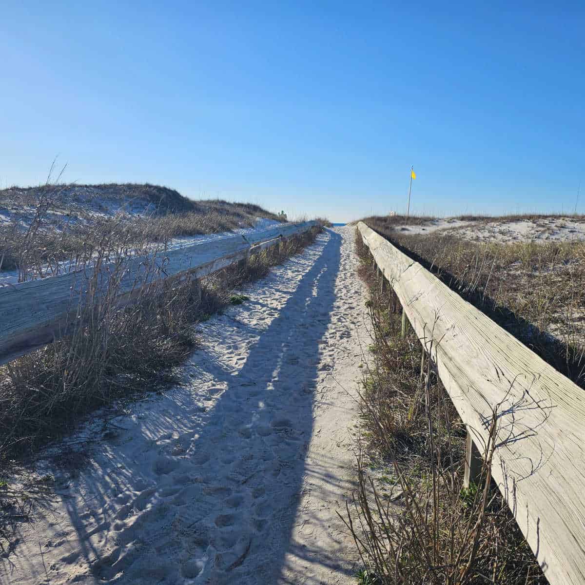 sand trail with wooden rails along the side leading to the beach with a yellow flag on a blue sky day