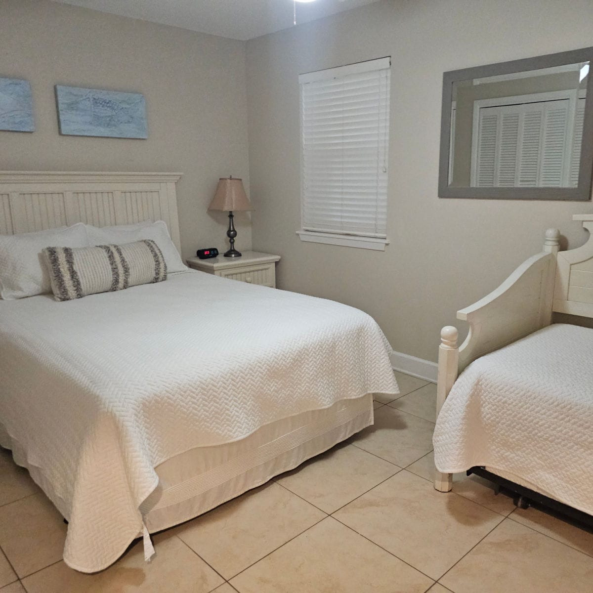 Bedroom with a queen bed with white linens and a striped pillow, a day bed with white linens, blue prints on the wall, nightstand with lamp and clock, mirror on the wall