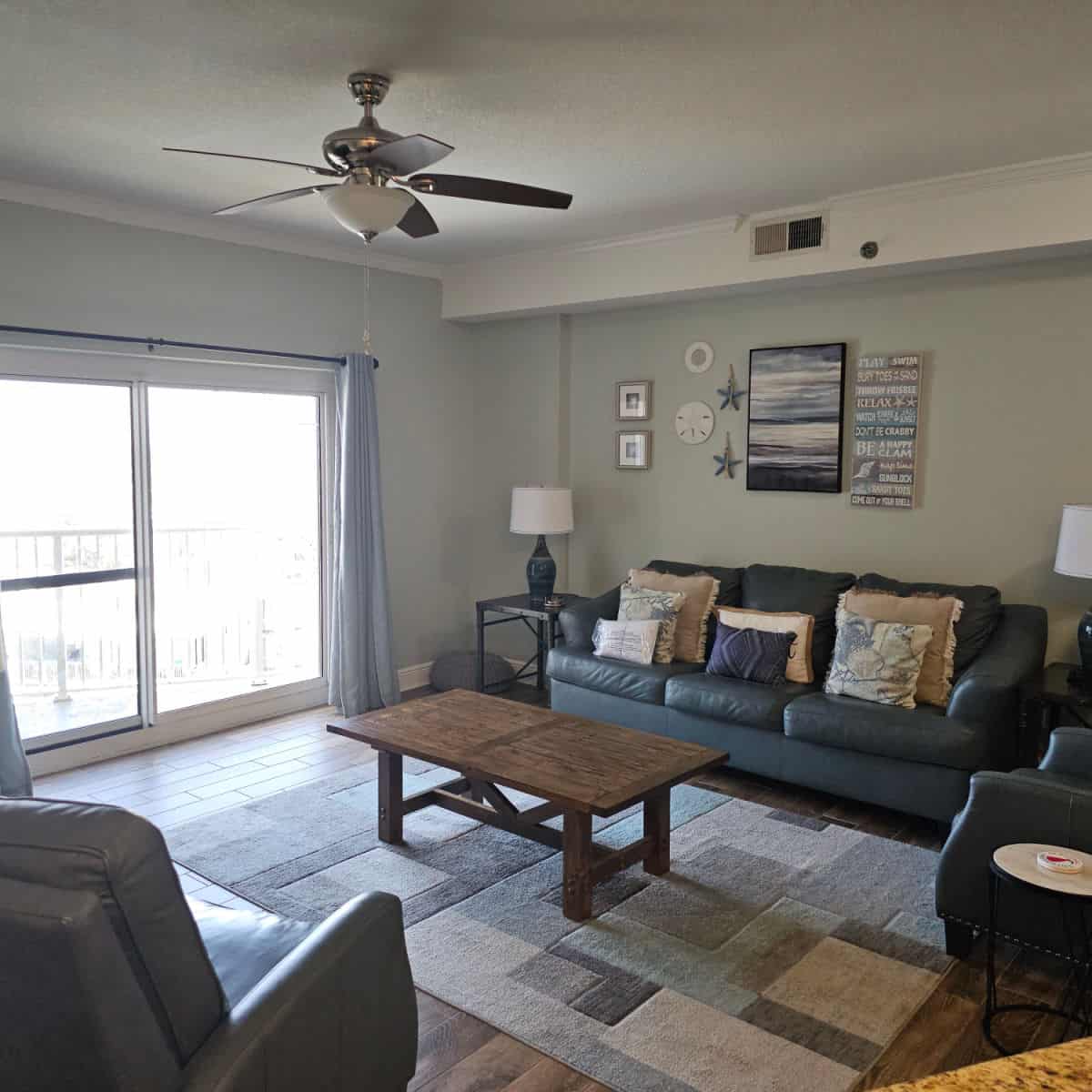 Living room with blue leather couch and chair, coastal art on the wall, table on a rug in the middle of the room, window looking out to the balcony