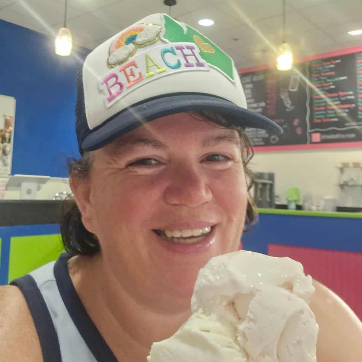 Tammilee wearing a beach hat holding an ice cream cone with a chalkboard menu behind her