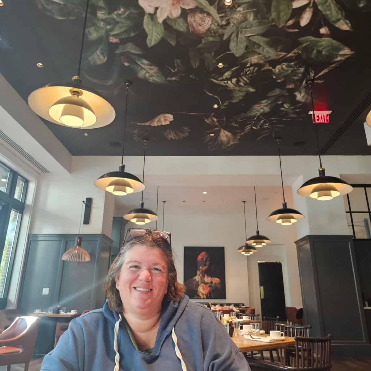 Tammilee smiling in a restaurant with lights overhead, floral paingint on the ceiling