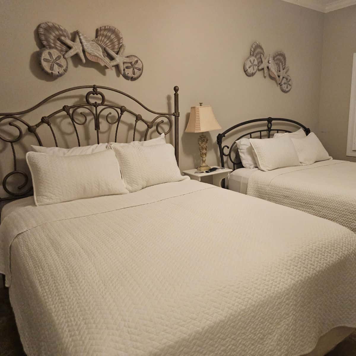 bedroom with two beds with wrought iron bed frames, shell art above the beds on the wall. Nightstand with a cream colored lamp